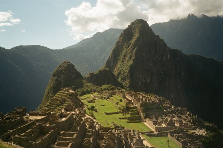 Machu Picchu in golden afternoon light — the Inca citadel on its ridge with Huayna Picchu behind, terraces and stonework tumbling down the green saddle.