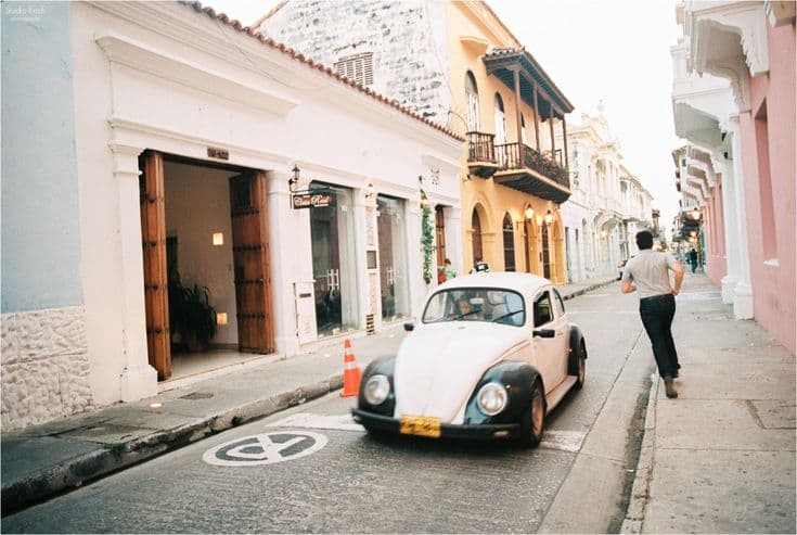 A street in Cartagena's walled old town at golden hour — a white VW Beetle on the cobbles, balconied colonial façades in ochre and pink.